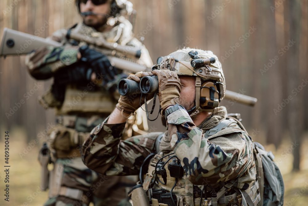 Portrait of a bearded middle-aged soldier in a Woodland military uniform and a helmet with headphones on his head, holding a rifle and looking around through the thick pine woods.